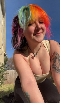 Curvy woman with multicolored hair smiles in beige tank top and black pants outdoors by fence.