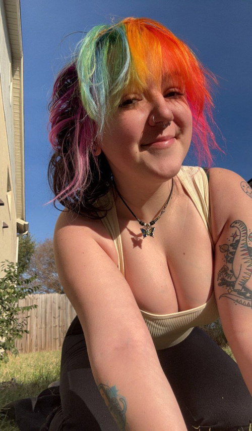 Woman with multicolored hair smiles outdoors wearing low-cut beige tank top with cleavage, black pants, arm tattoos,