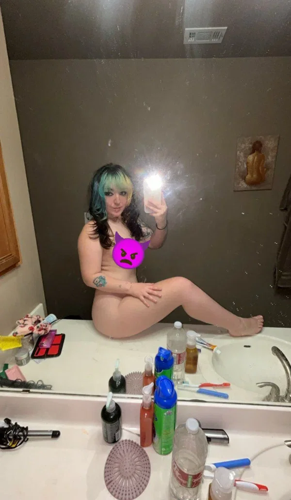 A woman with blue and black hair sits nude on a bathroom counter, taking a mirror selfie with her phone; tattoos are