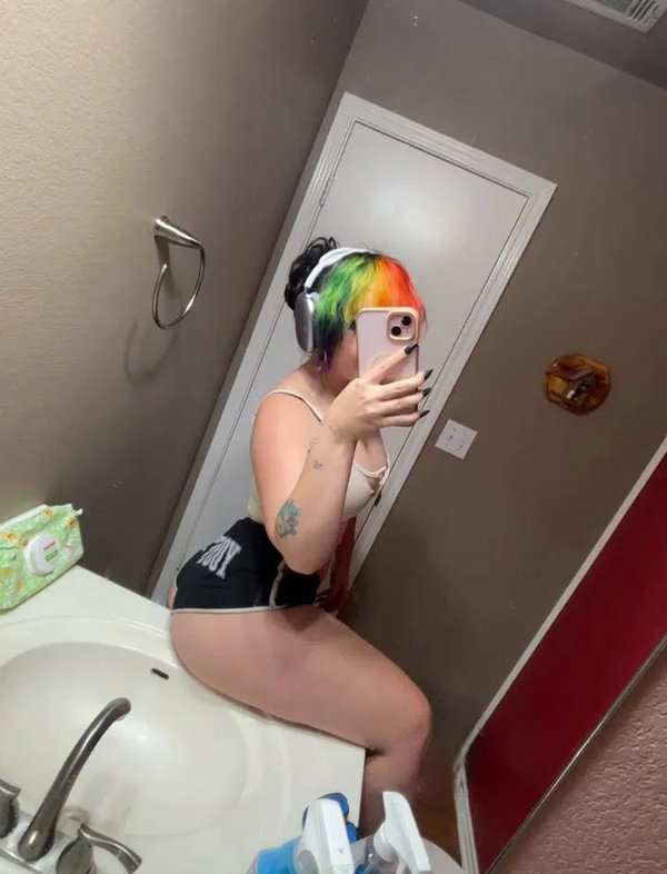 A person with rainbow hair and headphones sits on a bathroom sink, wearing a white top and black shorts, taking a