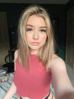 Blonde woman in a pink top sitting on a bed in a bedroom.