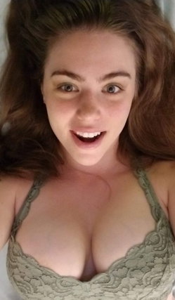 A woman with wavy brown hair smiles at the camera, wearing a green lace bra in a close-up selfie.