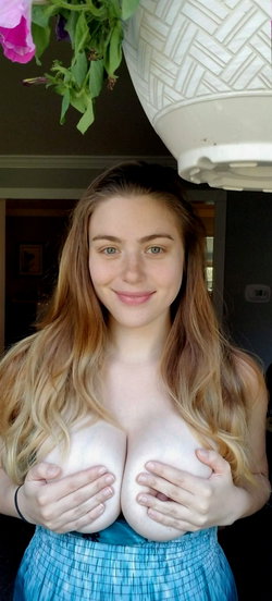 A woman with long wavy hair and green eyes smiles at the camera, holding her bare breasts while wearing a blue