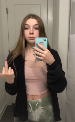 A young woman with long light brown hair takes a mirror selfie in a bathroom, wearing a black jacket over a pink crop