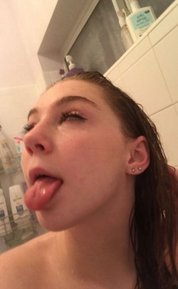 A young woman with wet dark hair sticks her tongue out in a bathroom selfie, bare shoulders visible.