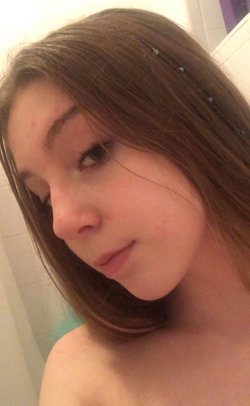 Close-up selfie of a woman with shoulder-length brown hair, bare shoulders, and a soft smile in a tiled bathroom.