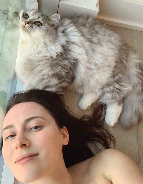 Brunette woman lying on her back, bare shoulders visible, with a fluffy gray cat resting on her head near a window.