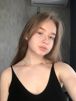 A young woman with light brown hair and blue eyes wears a black tank top, posing indoors near a wall with patterned