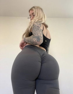 Blonde woman with full tattoo sleeve poses in black crop top and gray leggings, showing curvy figure against plain wall.