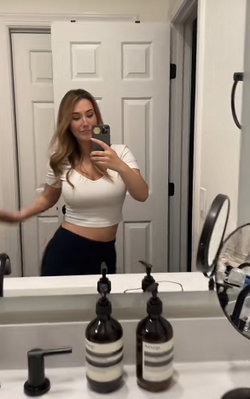 Woman with blonde hair takes mirror selfie in bathroom, wearing white crop top and dark bottoms.
