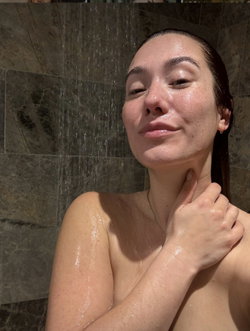 Woman with dark hair under shower, skin glistening, hand on neck, stone tile background.