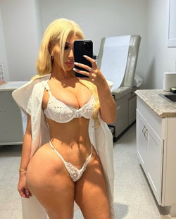 Blonde woman in white lace lingerie taking a selfie in a medical examination room.