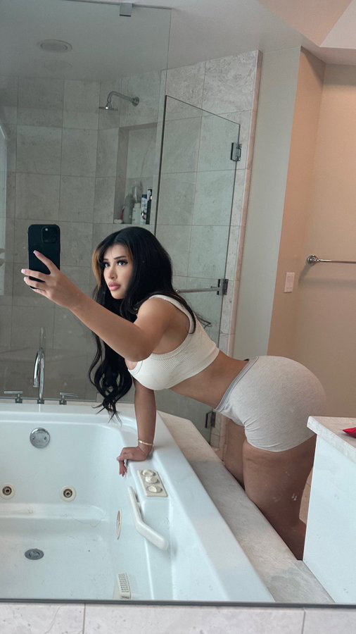 A curvy woman with long dark hair takes a mirror selfie in a bathroom, wearing a white crop top and matching shorts,