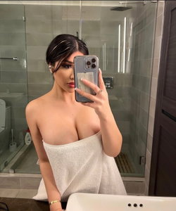Woman with dark hair wrapped in a white towel takes a mirror selfie in a bathroom.