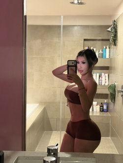 A woman with dark hair in a bun takes a mirror selfie in a bathroom, wearing a brown bandeau top and matching shorts,