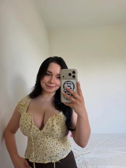 Curvy woman with long dark hair takes a mirror selfie in a floral top and brown pants, standing near a bed in a bright