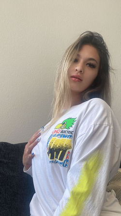 A woman with long ombre hair wears an oversized graphic tee, posing indoors with one hand on her chest.