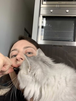 A woman with dark hair holds a fluffy gray cat close to her face in a kitchen, taking a selfie with a stainless steel