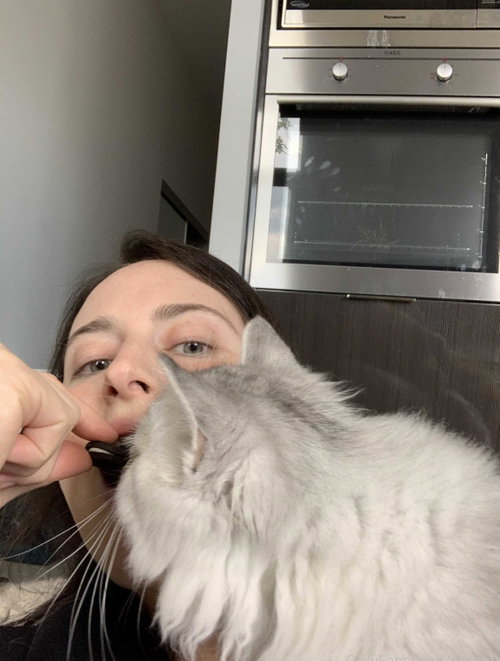 A woman with dark hair holds a fluffy gray cat close to her face in a kitchen, taking a selfie with a stainless steel