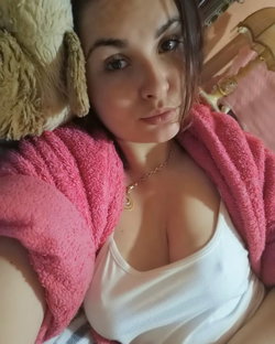 Brunette woman in a white tank top and pink robe, lying down with a plush toy, looking at the camera.