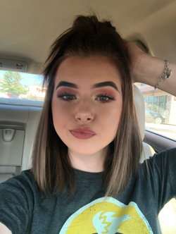 Brunette woman with makeup taking a selfie in a car, wearing a gray graphic tee and holding her hair up.