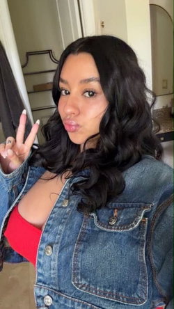 Brunette woman with wavy hair wearing a denim jacket over a red top, making a peace sign in a bathroom setting.