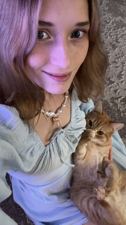 A young woman with purple-tinted hair smiles while holding a ginger cat, wearing a light blue ruffled top and a pearl
