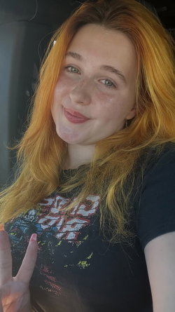 Redheaded woman with freckles smiles in a car selfie, wearing a graphic tee and making a peace sign.