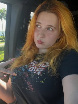 Redheaded woman with freckles and a graphic tee poses playfully in a car, pointing to her chest with a pouty expression.
