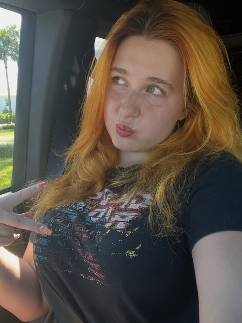 Redheaded woman with freckles and a graphic tee poses playfully in a car, pointing to her chest with a pouty expression.
