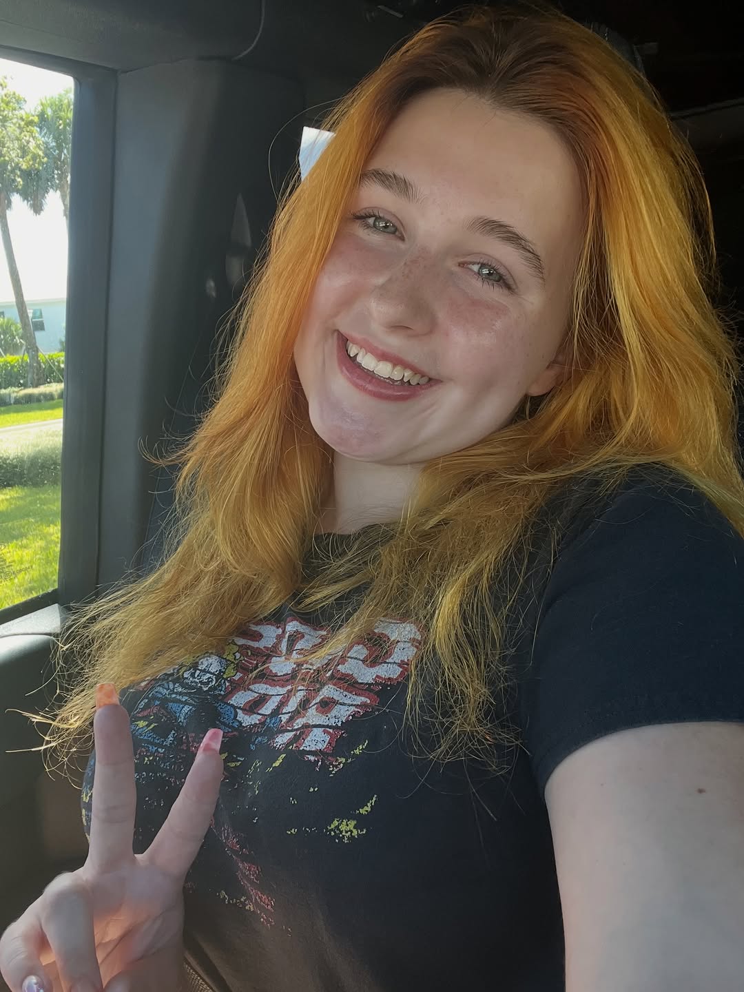 Redheaded woman with freckles smiling in a car, wearing a graphic tee and making a peace sign.