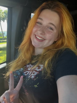 Redheaded woman with freckles smiling in a car, wearing a graphic tee and making a peace sign.