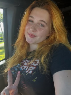 Redheaded woman with freckles smiling in a car, wearing a graphic tee and making a peace sign.