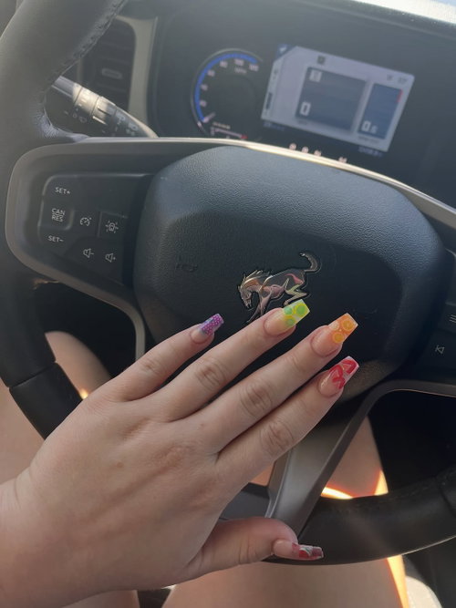 A hand with colorful nail art rests on a Mustang steering wheel inside a car, showing legs in light-colored shorts.