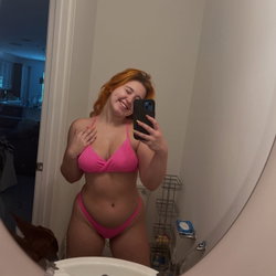 A woman with red hair in a pink bikini takes a mirror selfie in a bathroom, smiling with one hand on her chest.