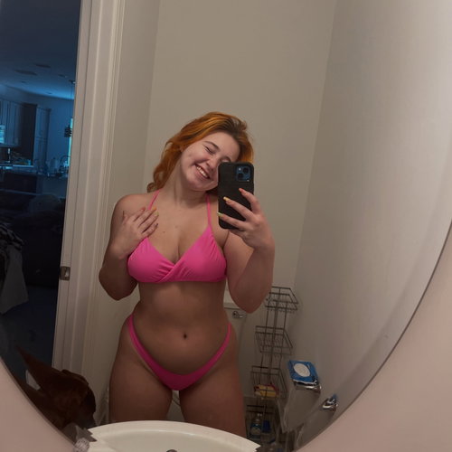 A woman with red hair in a pink bikini takes a mirror selfie in a bathroom, smiling with one hand on her chest.