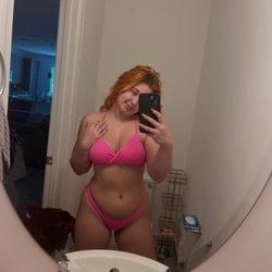 A woman with red hair in a pink bikini takes a mirror selfie in a bathroom, smiling softly.
