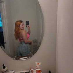 A woman with long red hair takes a mirror selfie in a pink top and denim shorts, smiling in a bathroom setting.