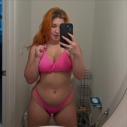 Redheaded woman in pink bikini takes mirror selfie in bathroom, holding phone with visible arm and hand.