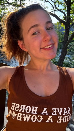 A smiling woman with brown hair in a messy bun wears a brown tank top outdoors near trees and water, taking a selfie.
