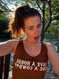 A young woman with brown hair in a high ponytail wears a brown tank top, making a playful face outdoors near a river