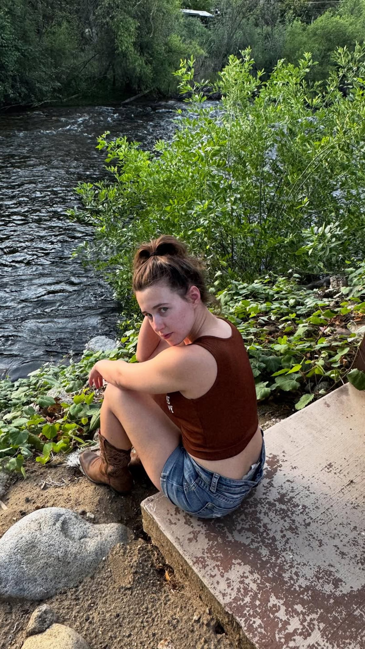 A woman with brown hair in a bun sits by a river, wearing a brown tank top and denim shorts, with cowboy boots,