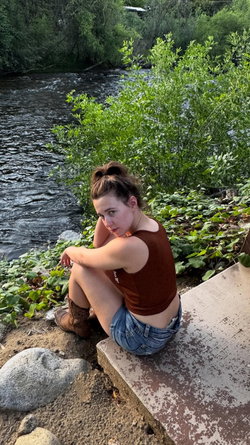 A woman with brown hair in a bun sits by a river, wearing a brown tank top and denim shorts, with cowboy boots,