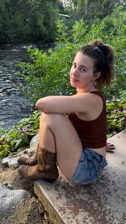 Curvy woman in a brown tank top and denim shorts sits by a river, wearing cowboy boots, with green foliage in the