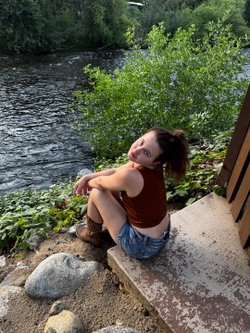 A woman with brown hair in a ponytail sits on a concrete step by a river, wearing a brown tank top and denim shorts,