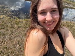 A smiling woman with long brown hair in a black tank top takes a selfie outdoors near a grassy pond.