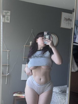 Curvy woman with long dark hair takes mirror selfie in gray crop top and white underwear, standing in bedroom with gray