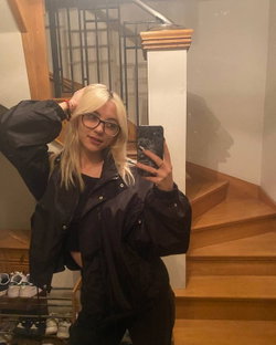 Slim blonde woman in glasses and open black jacket poses near wooden staircase.
