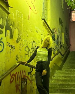 Slim blonde in cropped white top and black pants poses on yellow graffiti stairs.