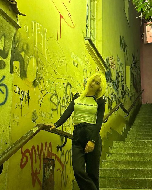 Slim blonde in cropped white top and black pants poses on yellow graffiti stairs.